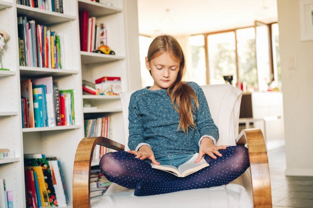 Cute little girl sitting in a white chair at home and reading a book